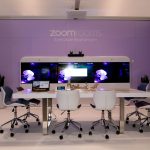 A Zoom Rooms executive boardroom with a large display, camera, microphone, table, chairs, and controller.