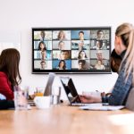 Zoom videoconferencing with 16 remote participants on the display and six in-person attendees at a table with laptops.