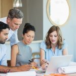 An office space with four employees sitting or standing around a desk with a laptop.