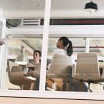 Exterior view of an office seen through glass walls. Inside the office, a man is standing and speaking to a seated man and woman.