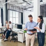 An office space with a man and a woman in the foreground, looking at a folder. Another man and woman are standing at desks in the background.