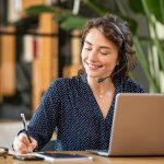A woman sitting in front of a laptop, wearing a headset with an earpiece and microphone.